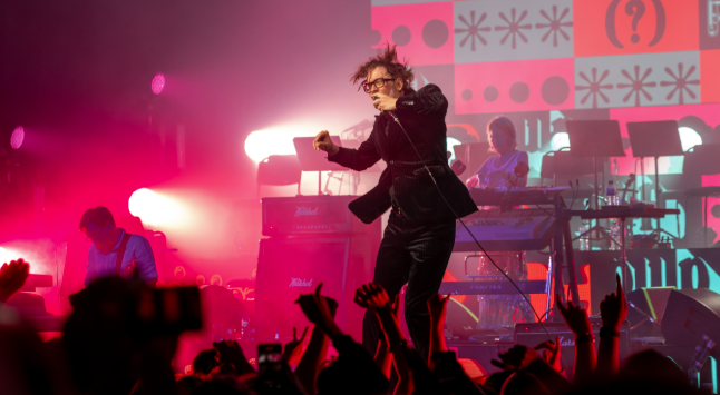 Jarvis Cocker performing live with Pulp under pink stage lights, arms raised in the crowd