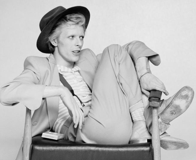 David Bowie reclining in a chair, wearing a light suit and hat