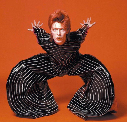 David Bowie posing against an orange background in a dramatic striped stage costume